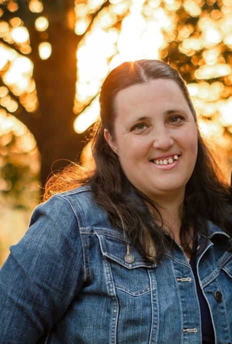 A woman in a denim jacket is smiling in front of a tree.