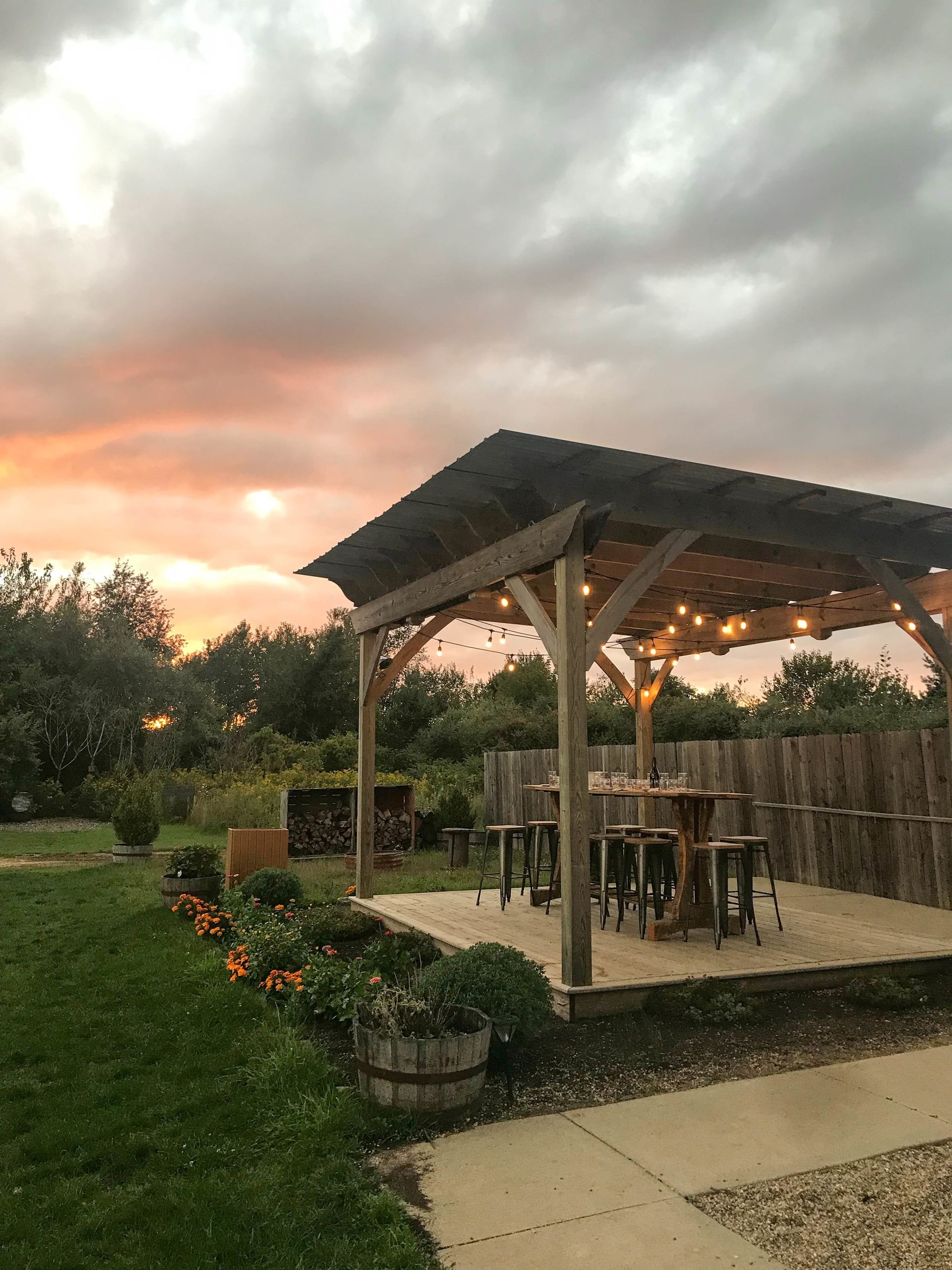 A wooden gazebo with tables and stools under a cloudy sky at sunset.