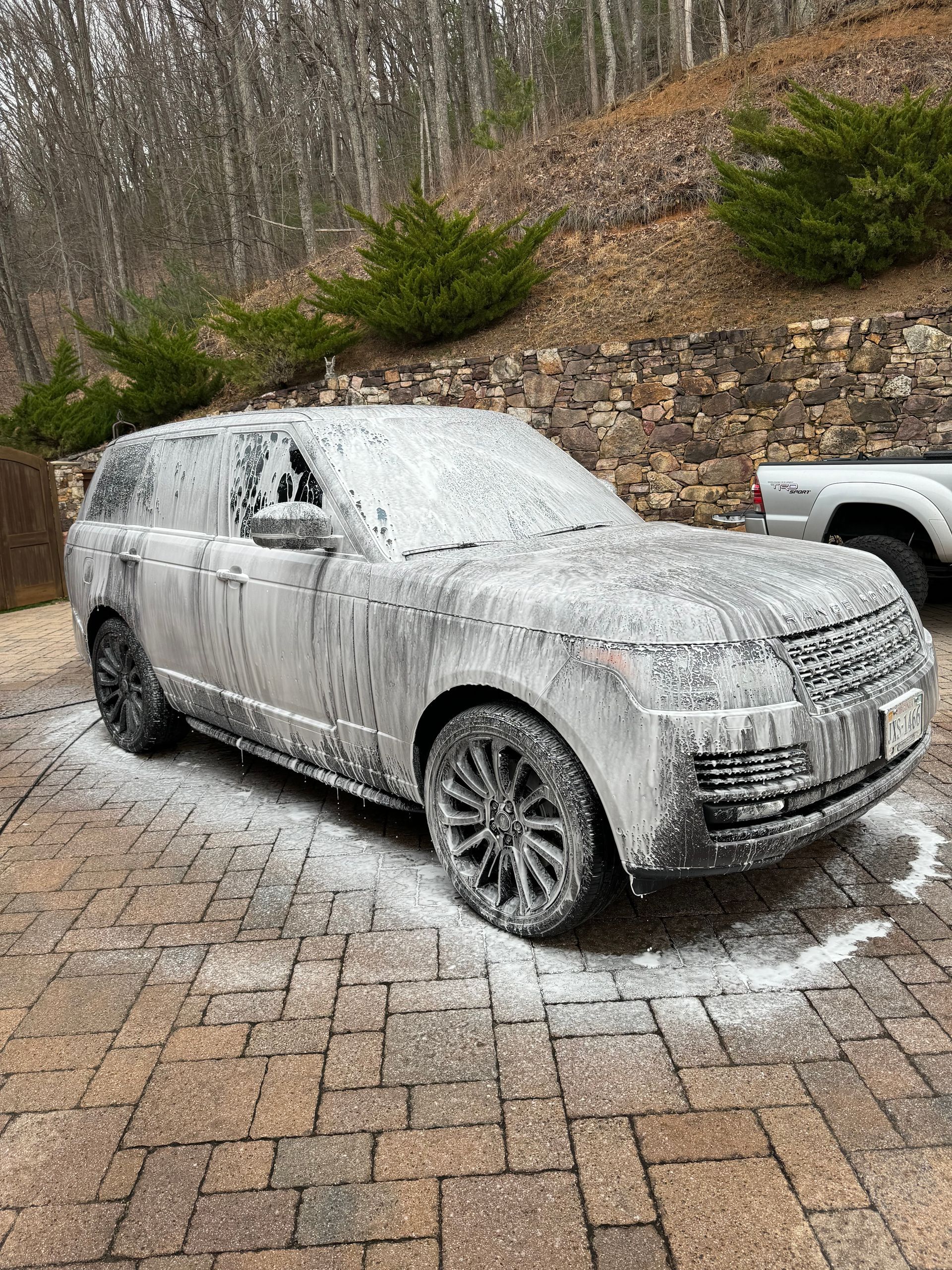 Mud-covered SUV parked on a brick driveway beside a wooded hillside, with snow on the ground.