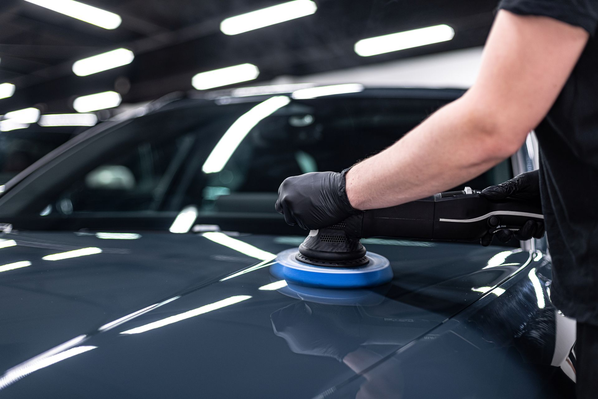 Person polishing a black car with a buffer in a garage-like setting