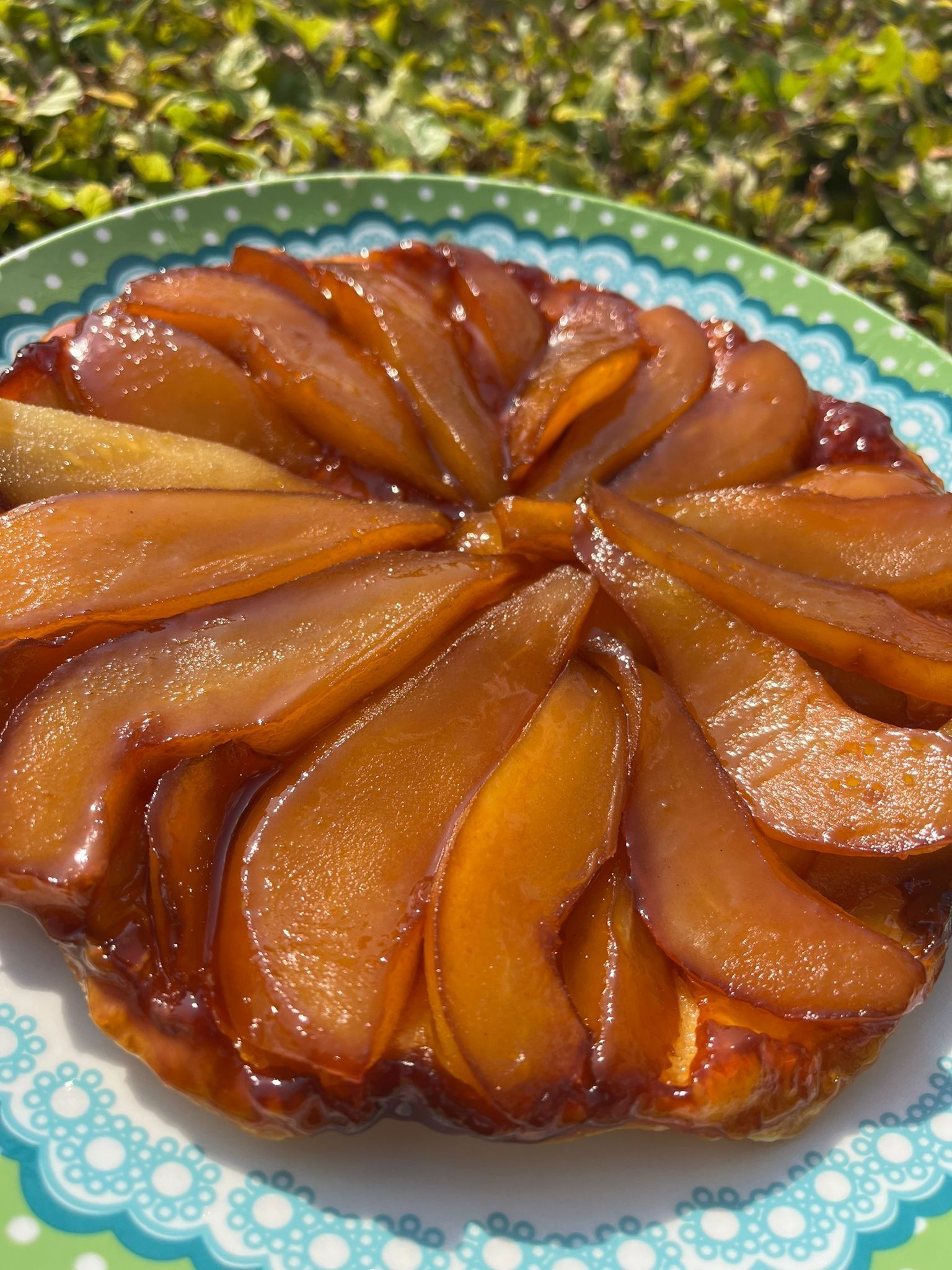 Een perentaart (tarte tatin) met goudbruine, gekarameliseerde fruitschijfjes in een cirkelvormig patroon op een bord met een groene rand.