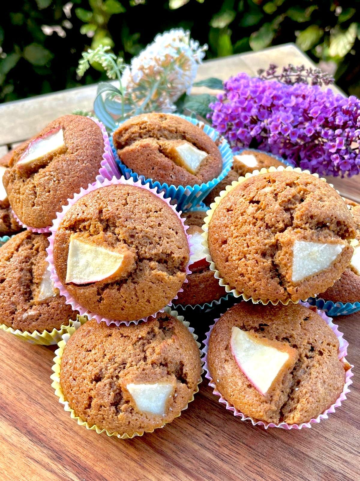 Een stapel goudbruine muffins, bedekt met plakjes peer, gepresenteerd in kleurrijke papieren vormpjes naast paarse bloemen.