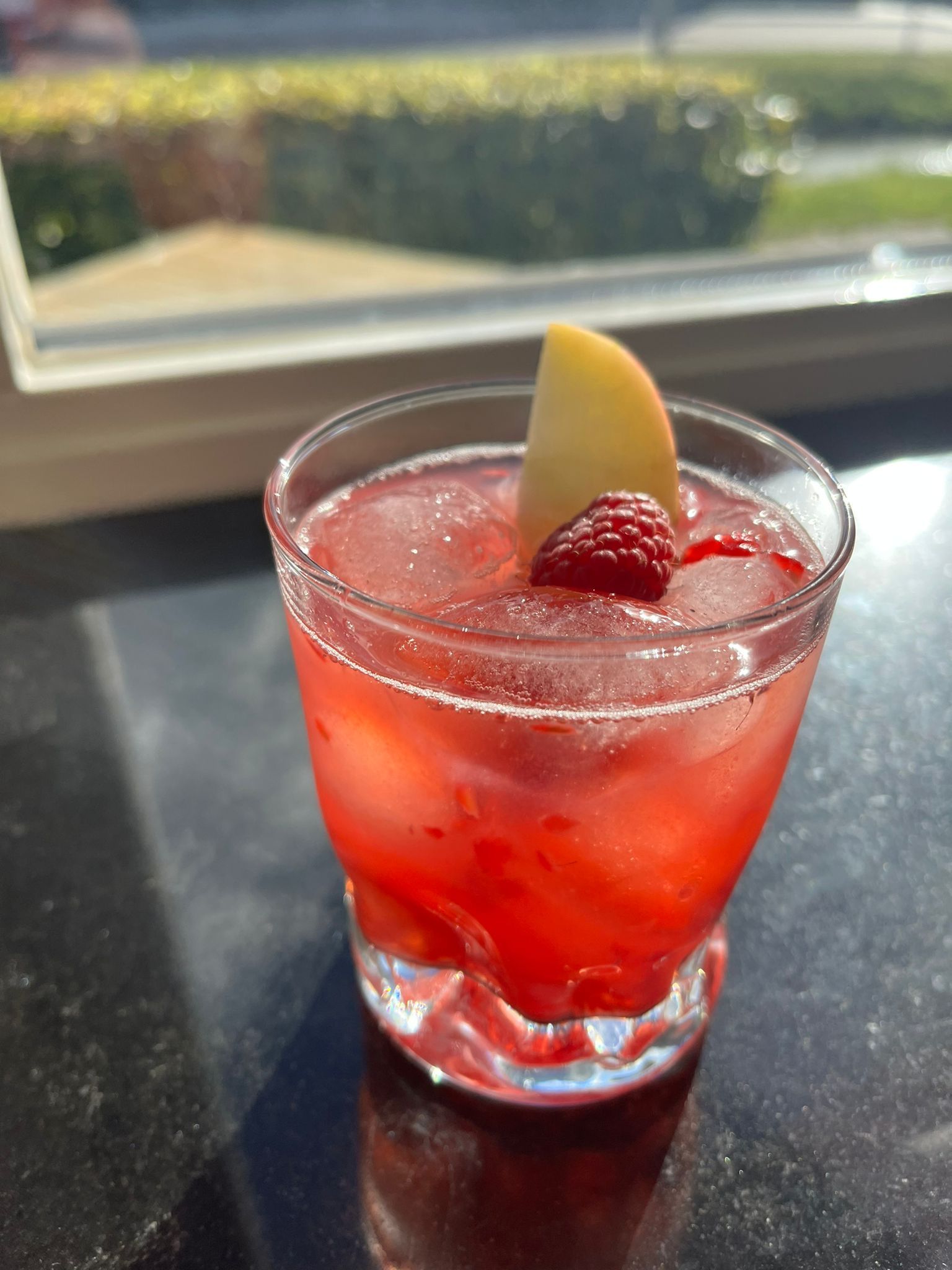 Een rode cocktail met ijs, gegarneerd met een verse framboos en een dun schijfje appel, staat op een donker oppervlak bij een raam.