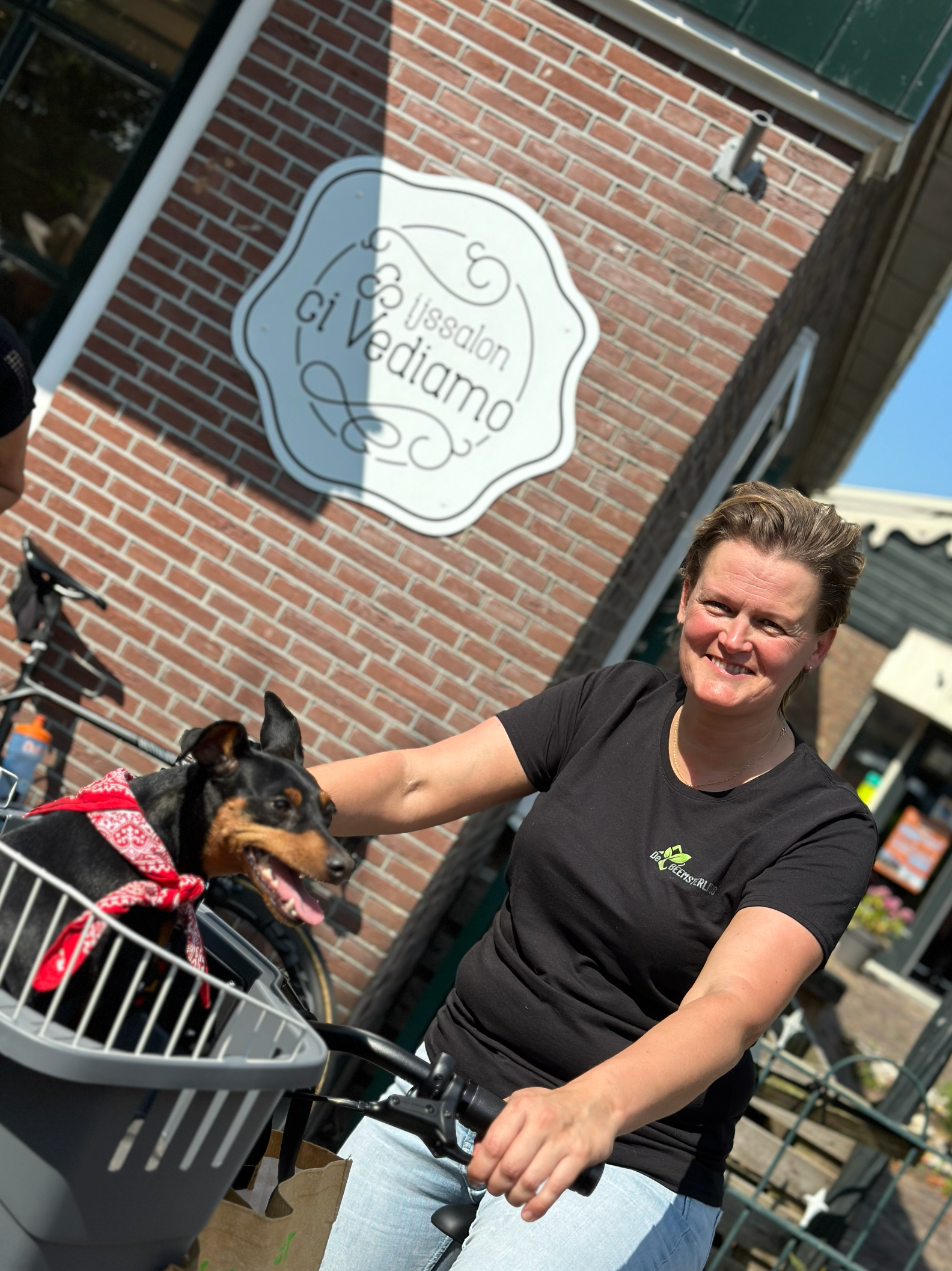 Een persoon lacht terwijl hij op een fiets rijdt met een hond in een mandje voorop, voor een bakstenen gebouw met een reclamebord.