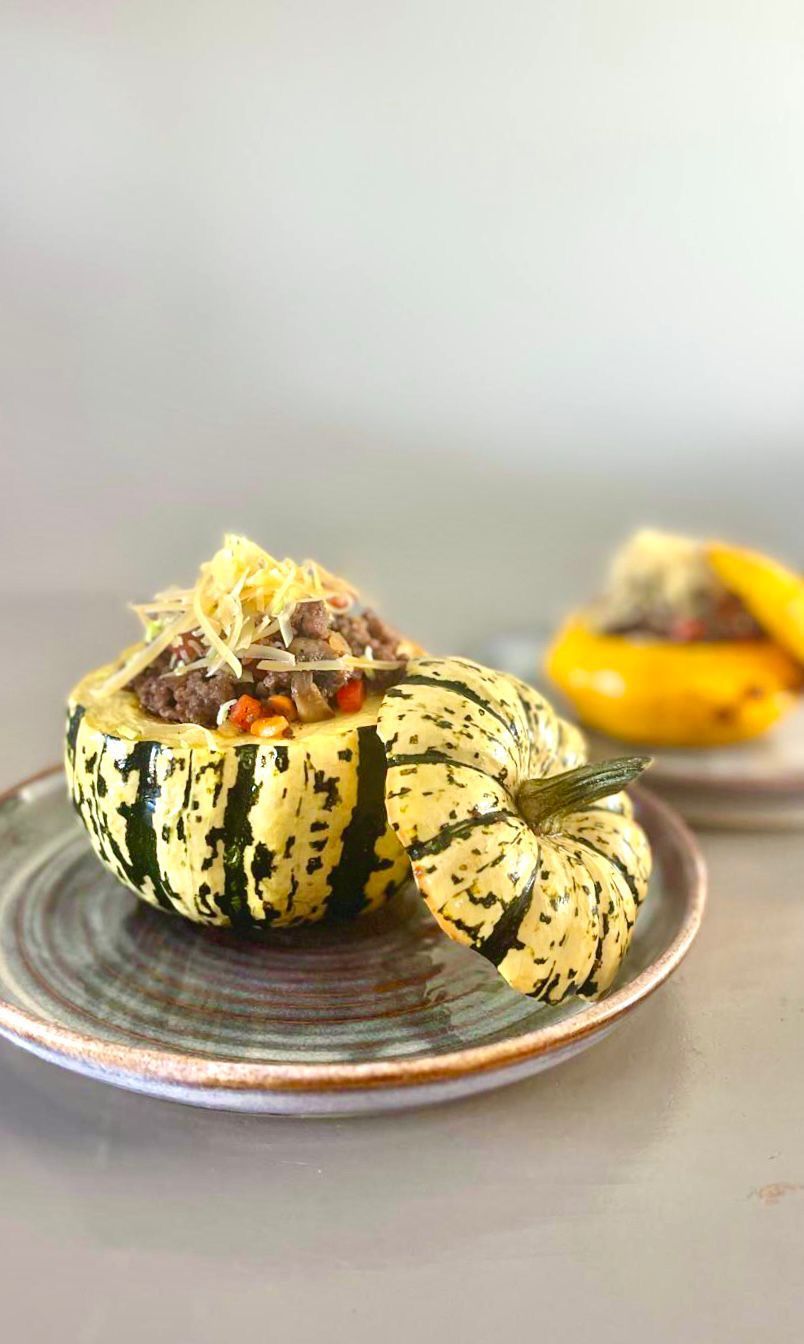 Twee gevulde, gestreepte delicata-pompoenhelften, bedekt met geraspte kaas, liggen op een grijs keramisch bord.