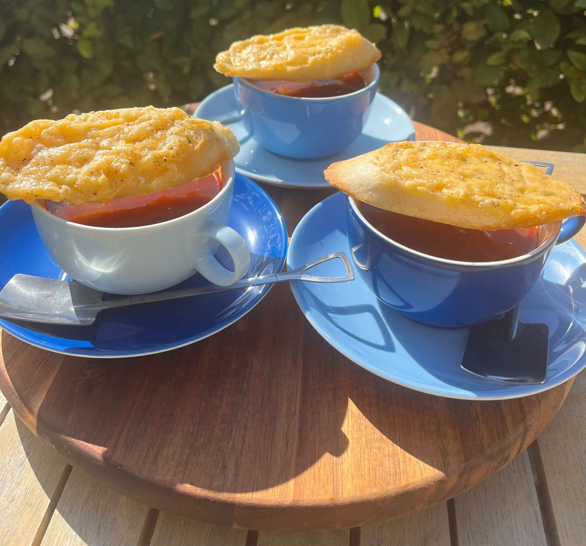 Drie kopjes tomatensoep, elk met een stukje geroosterd brood met kaas erop, geserveerd op blauwe schoteltjes op een houten plank.