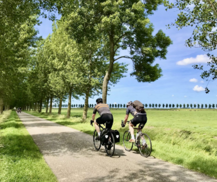 Twee fietsers rijden over een zonovergoten, geplaveid pad, omzoomd door hoge bomen, langs een uitgestrekt groen veld onder een helderblauwe hemel.