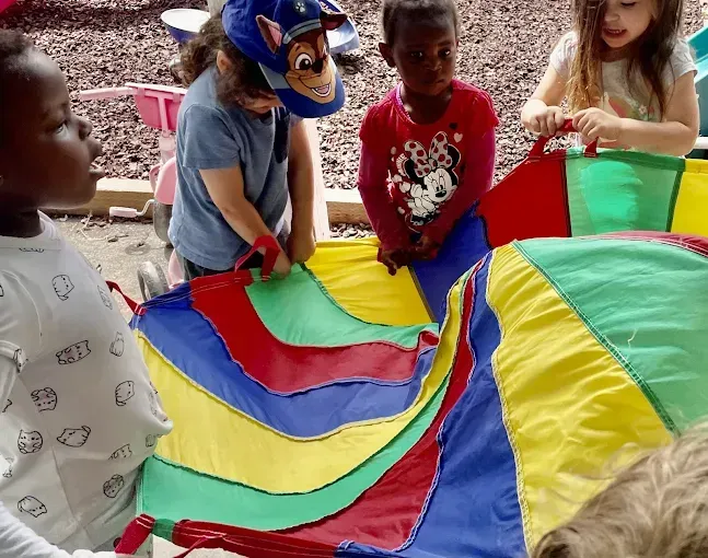 Children playing with a rainbow parachute outside; red, yellow, green, and blue fabric.