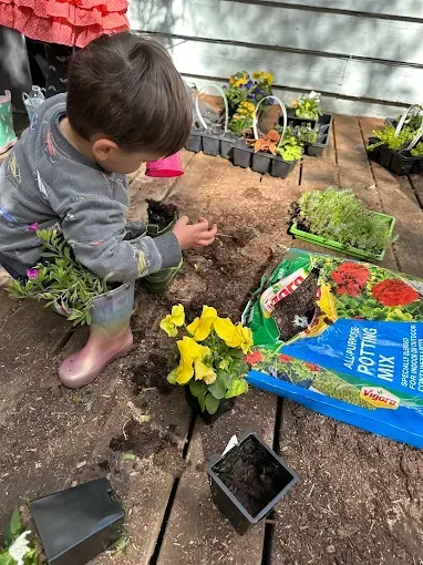 Young child planting flowers on a deck with potting mix and potted plants.