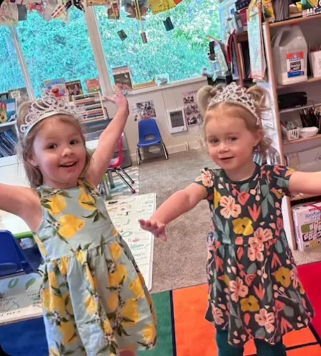 Two girls in tiaras and floral dresses, smiling and waving arms in a colorful classroom.