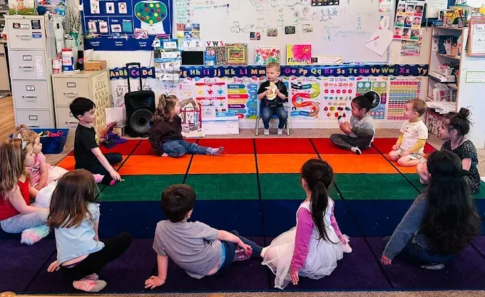 Children in a circle on a colorful rug, one child performs with a microphone in a classroom.