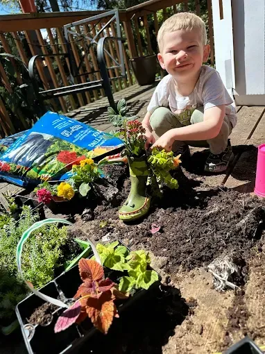 Boy planting flowers in a garden, smiling, with a green boot planter.