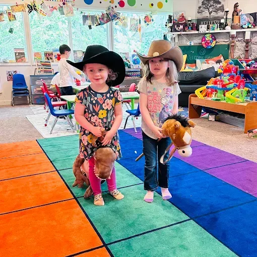 Two children wearing cowboy hats, riding stick horses on a colorful mat in a playroom.