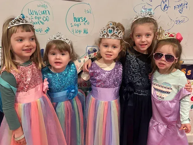 Five young children in princess costumes and tiaras, smiling. They stand in front of a whiteboard.