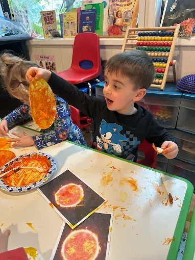 Boy holding up painted sun art, smiling. Sun art cards and paint visible on table.