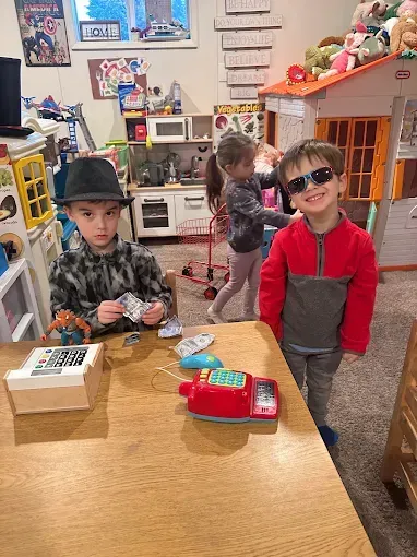 Three children playing store indoors, one wearing sunglasses and smiling, others near a play cash register.