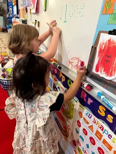 Two children drawing on a whiteboard with markers; one child is blond, and the other has dark hair.