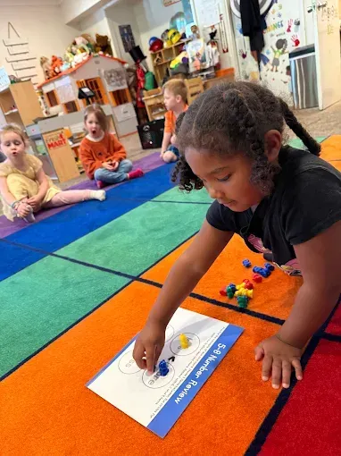 Girl playing with colorful blocks on a rug. Other children sit and watch in a classroom.