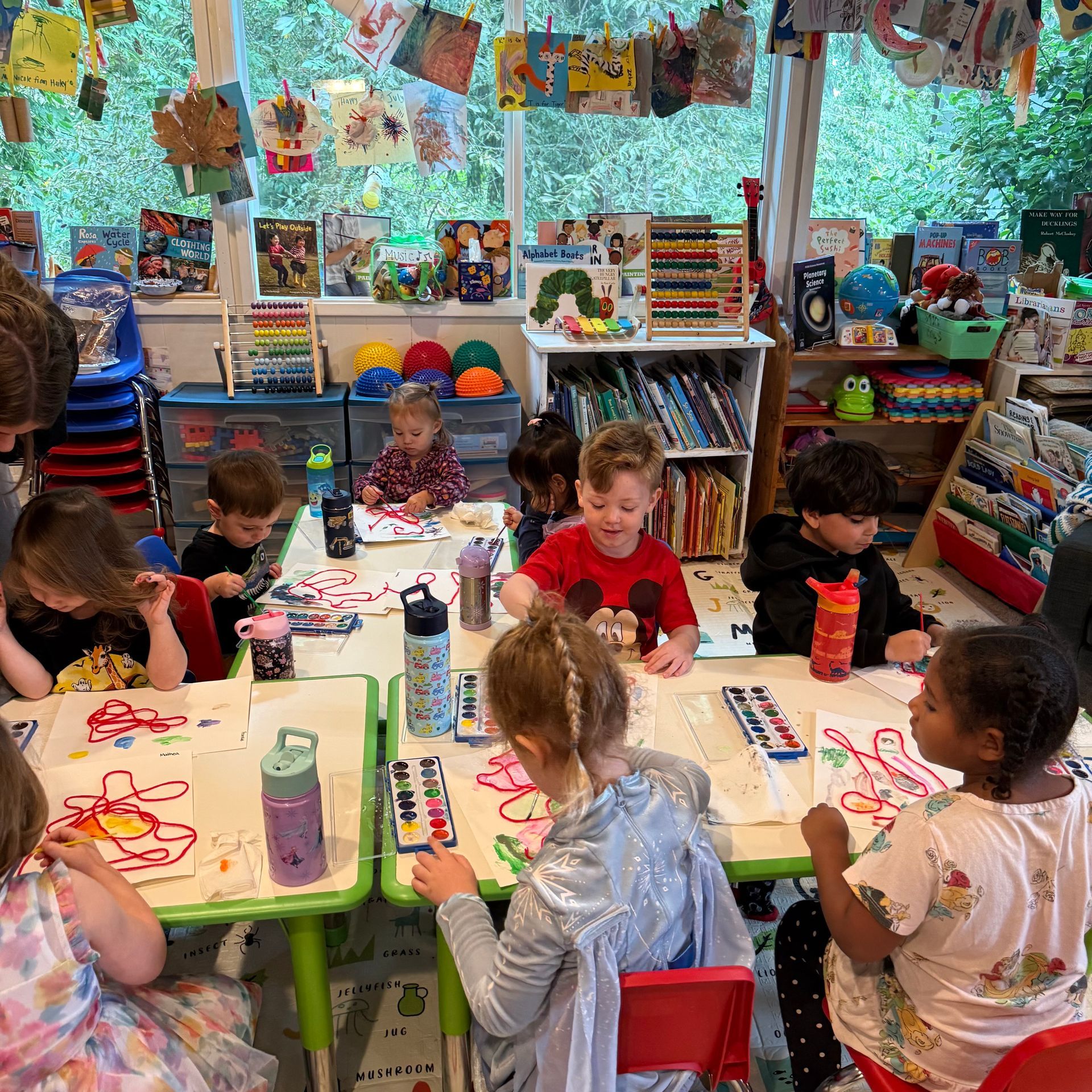 Children painting at tables in a brightly decorated classroom, surrounded by art supplies and books.