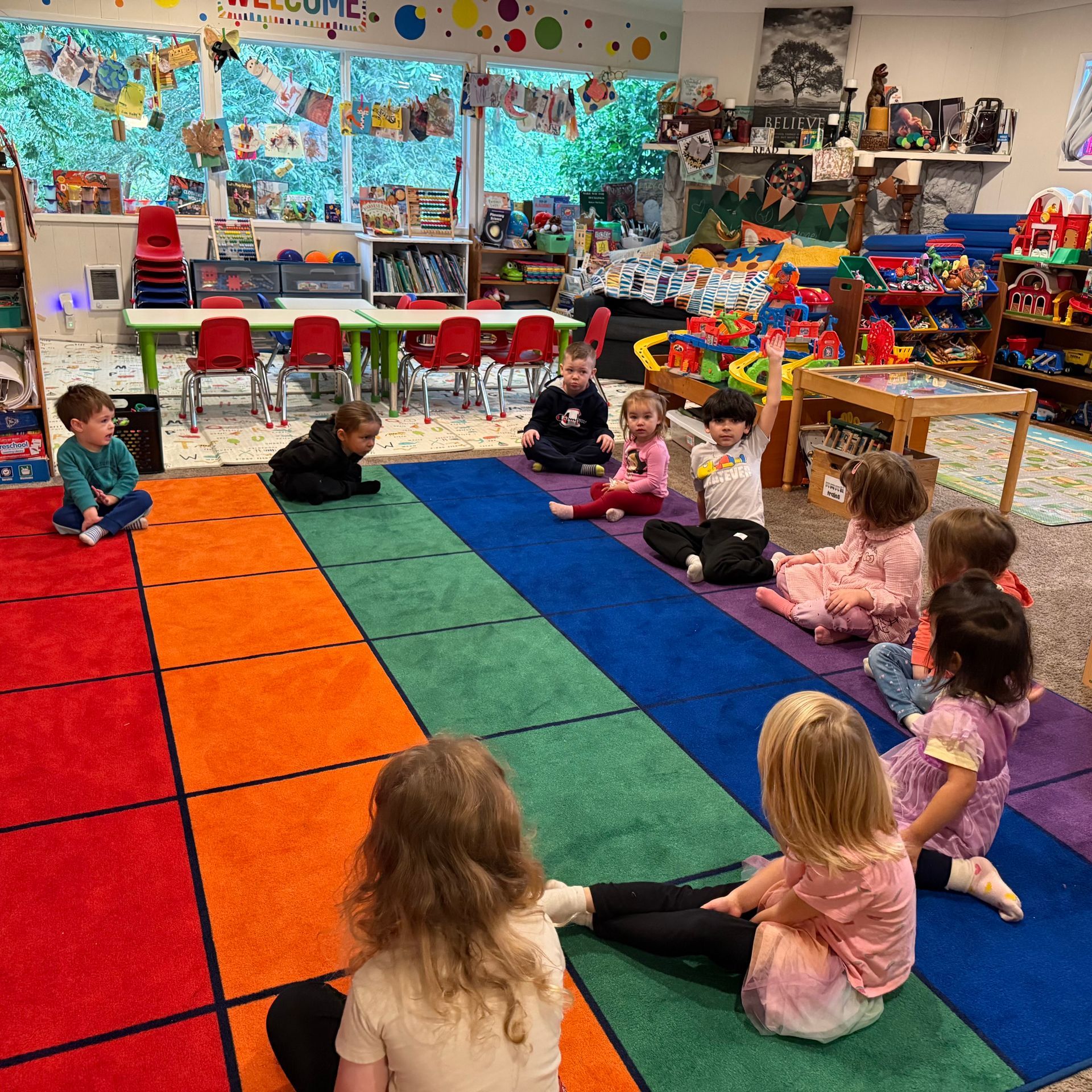 Children sitting in a circle on a rainbow rug in a colorful classroom.