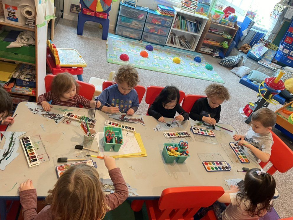 Children painting at a table in a classroom. They are using watercolors, with paint supplies and red chairs.
