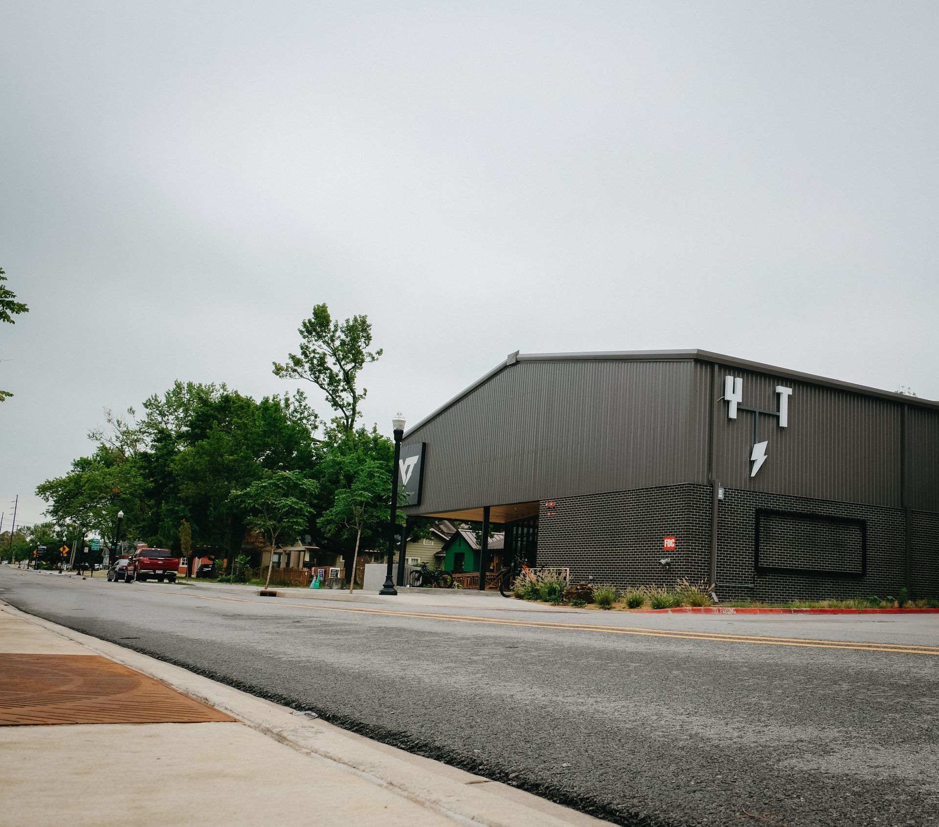 Asphalt street with a large gray metal building and green trees under a cloudy sky.