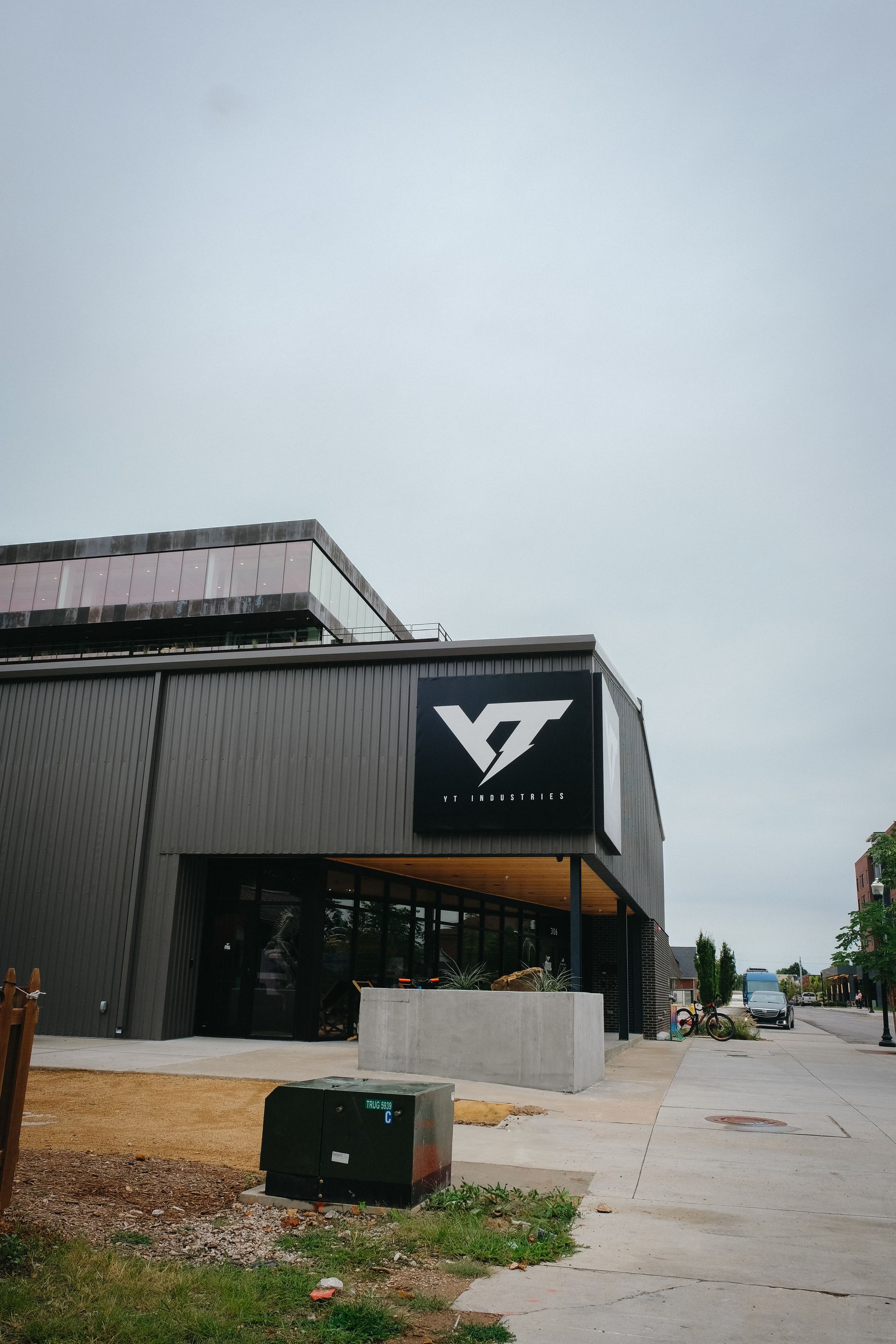 Modern building with a large logo, cloudy sky. Sidewalk and street with a few parked vehicles.