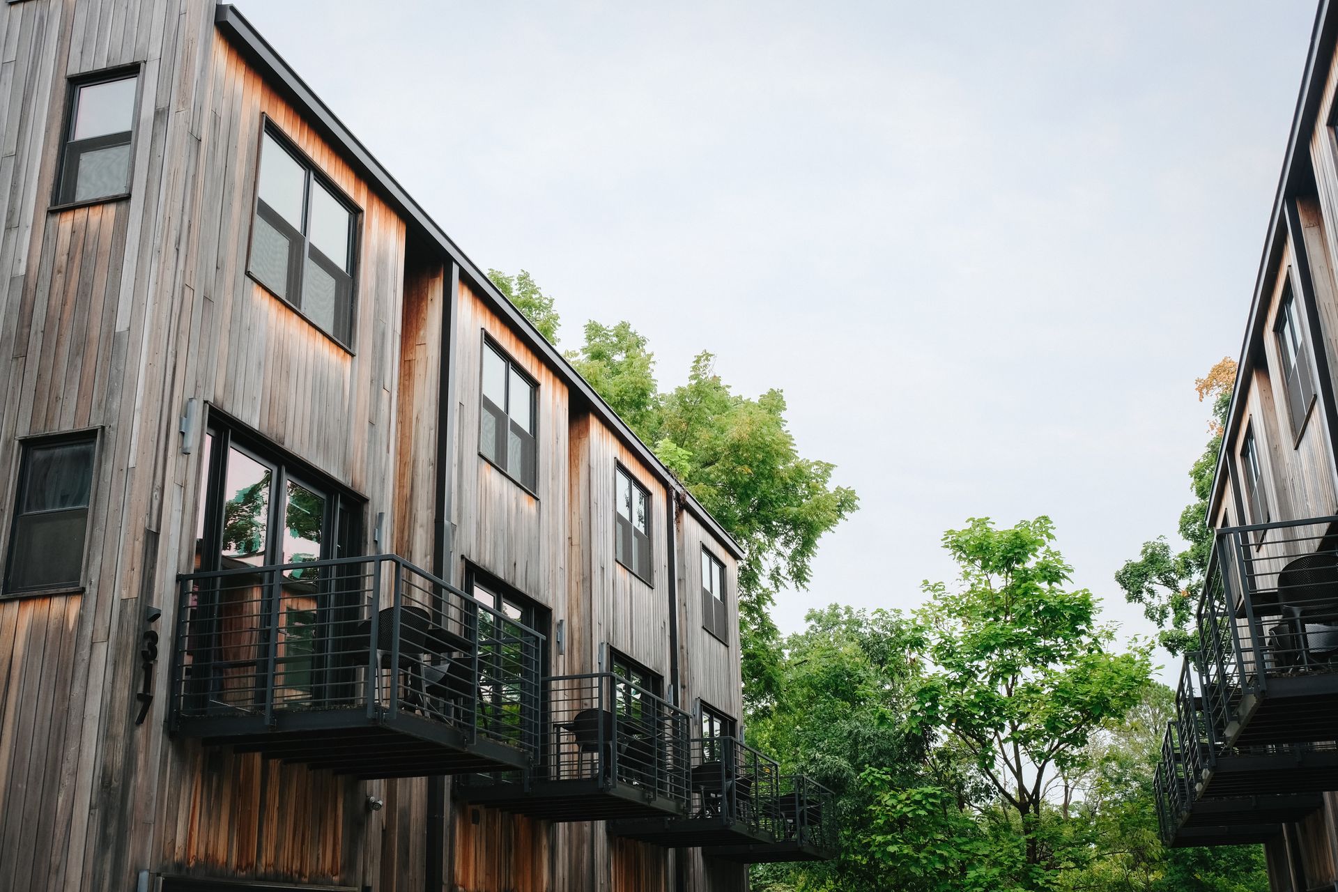 Two wooden apartment buildings with black window frames and balconies; green trees in the background.