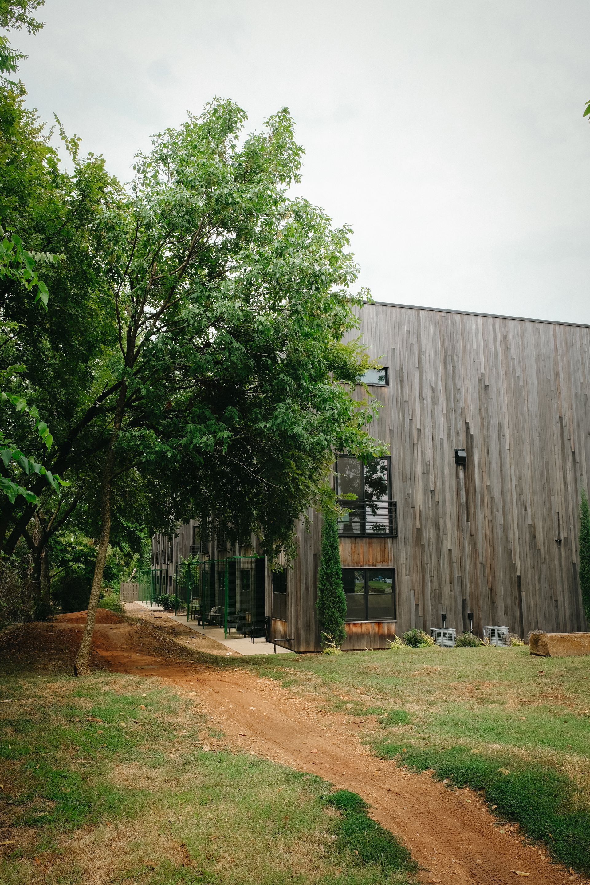 Wooden building with dark windows, set in a grassy area with a dirt path and trees.