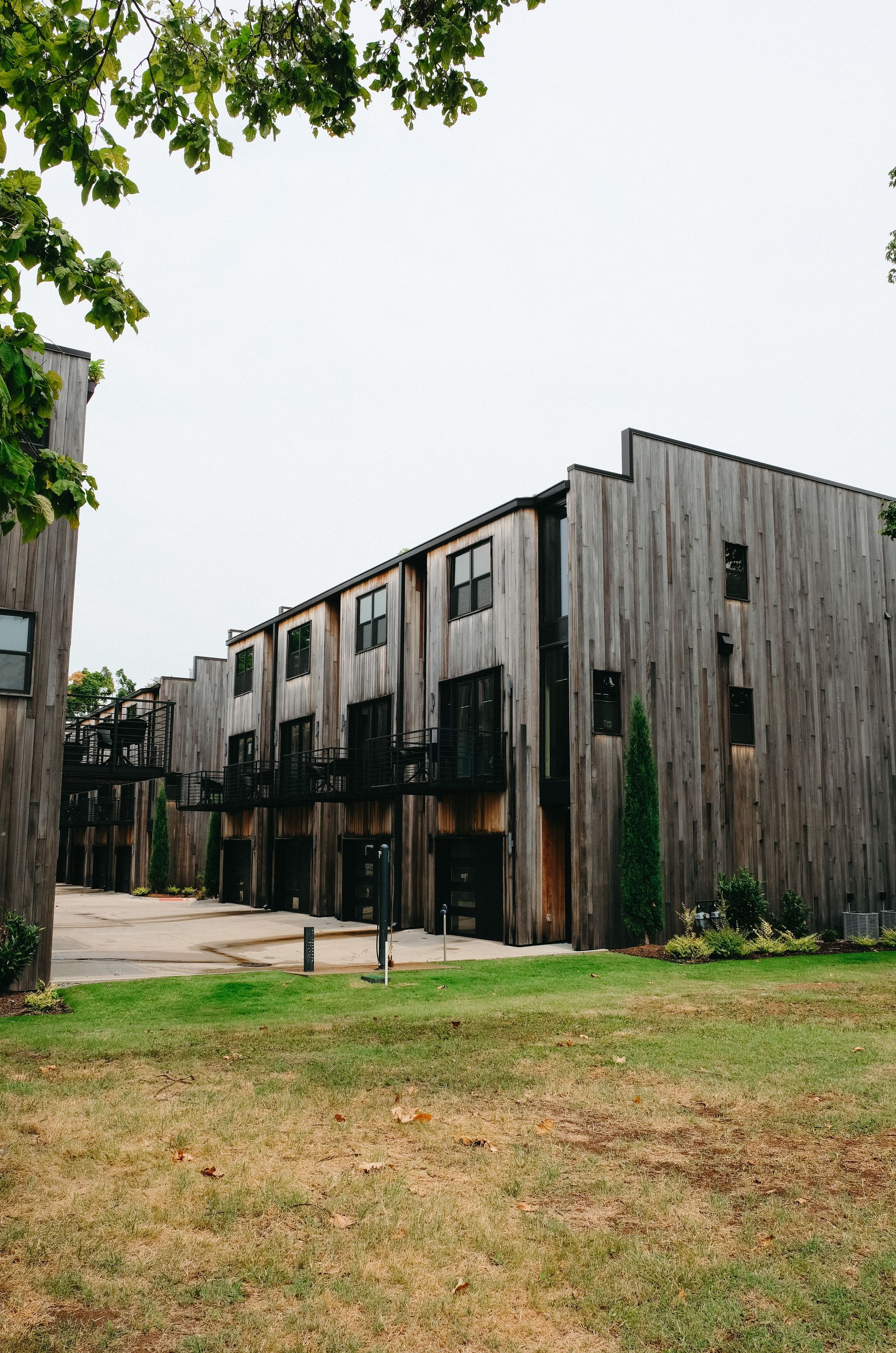 Wood-sided townhouses with black balconies and windows, set on a grassy lawn under a cloudy sky.