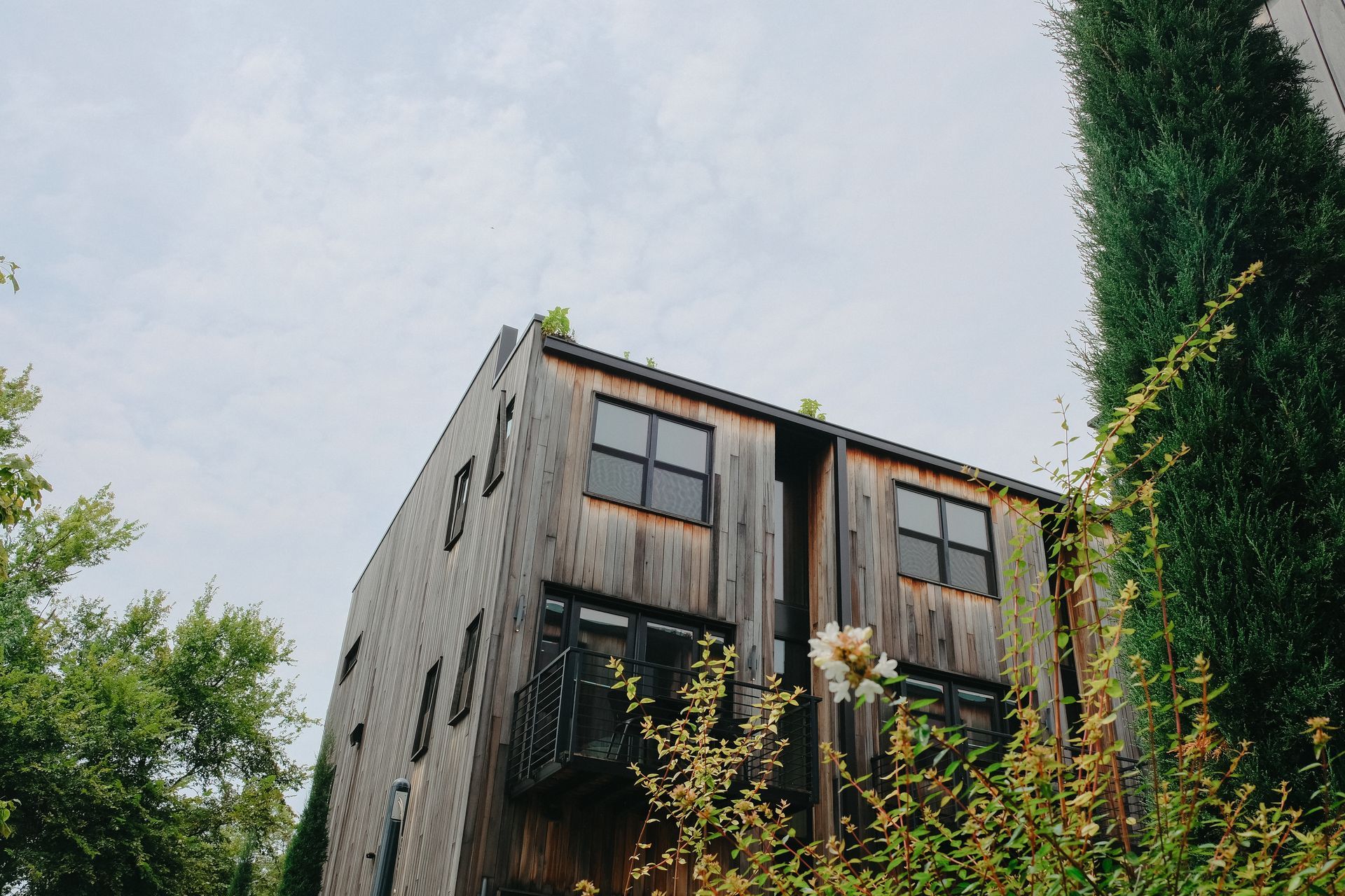 Wooden building with dark windows and lush greenery against cloudy sky.