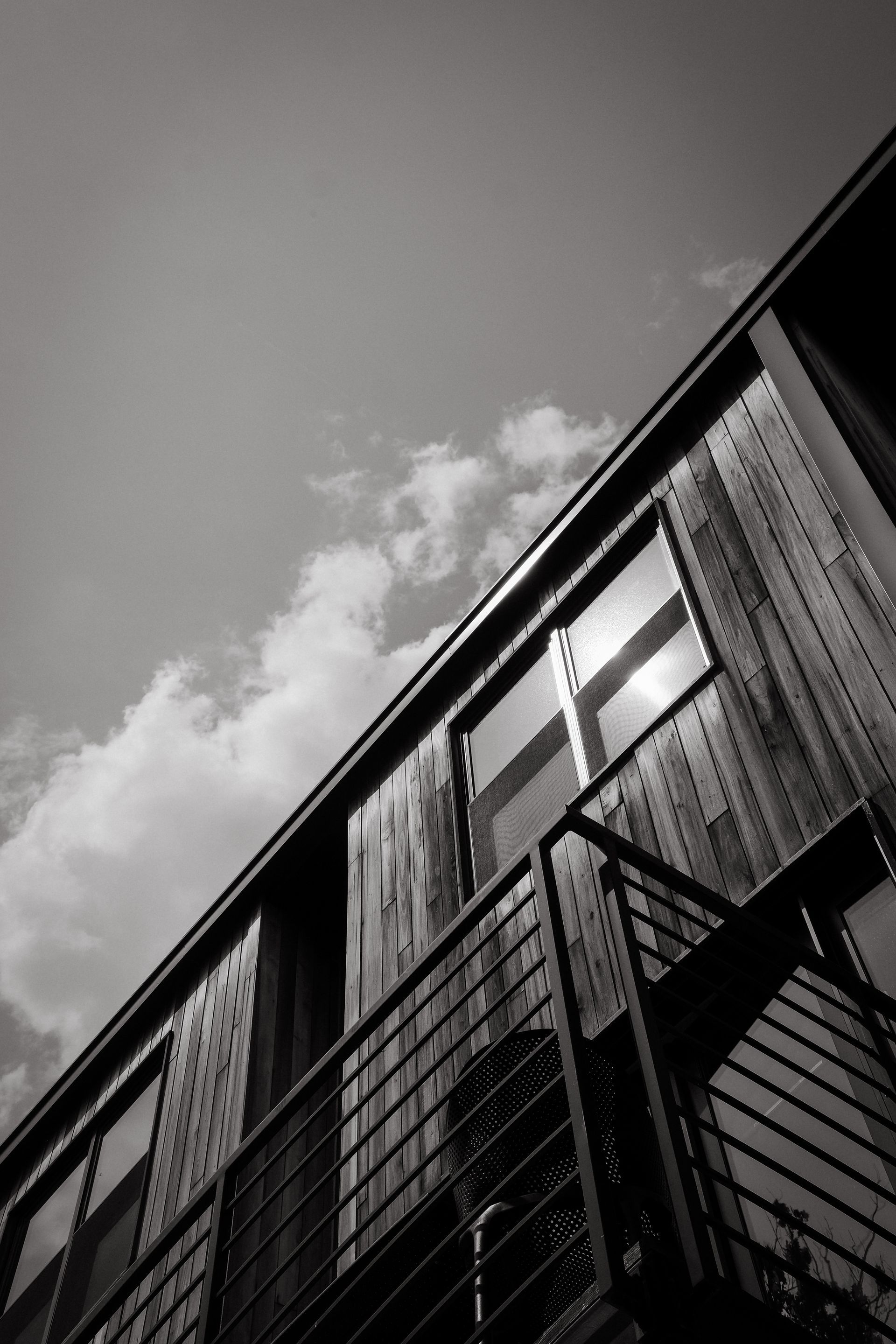 Black and white shot of a modern building with a balcony, windows, and wooden siding, angled upwards towards the sky.