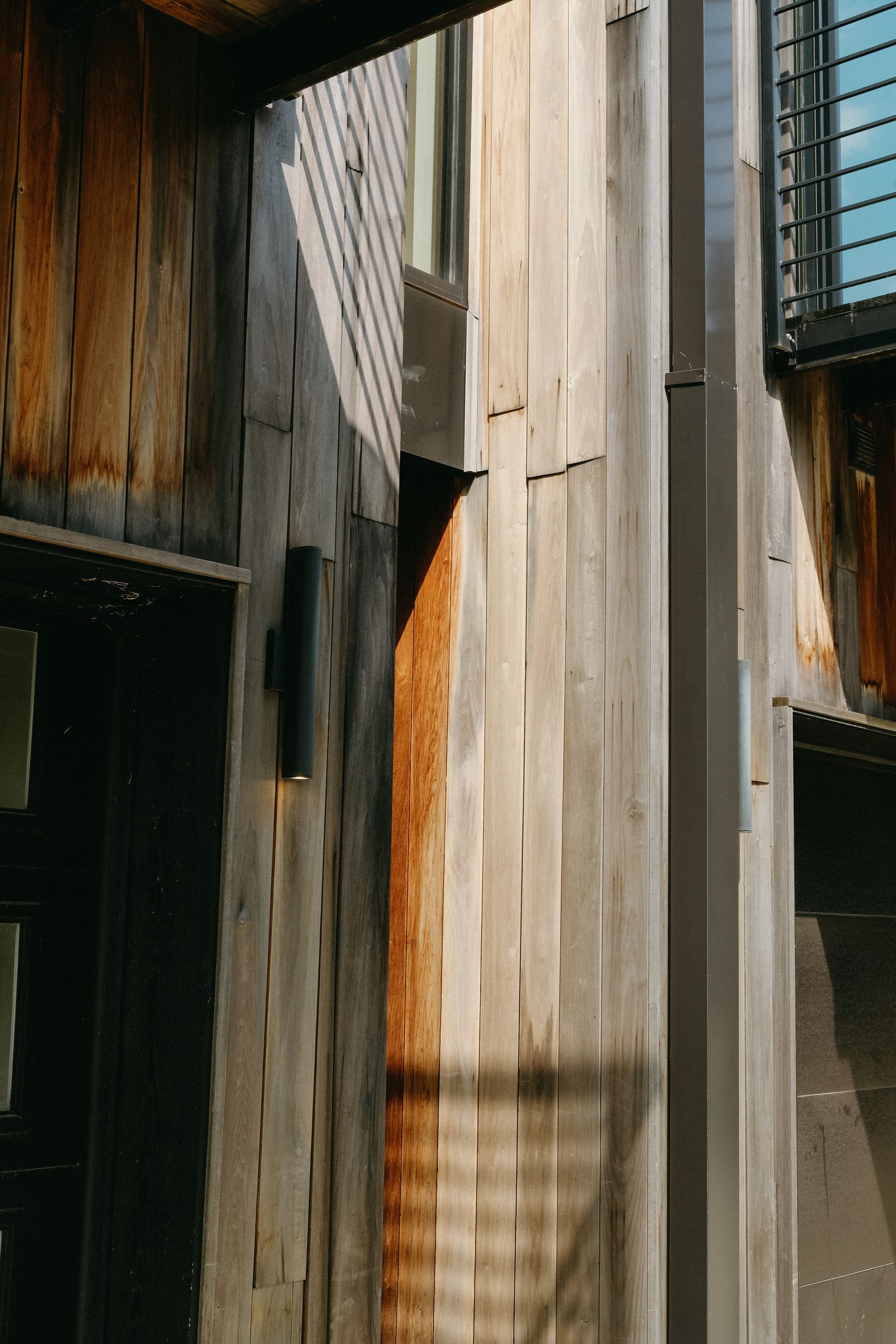 Weathered wood siding on a building, illuminated by sunlight. Dark accents, windows, and a modern sconce.
