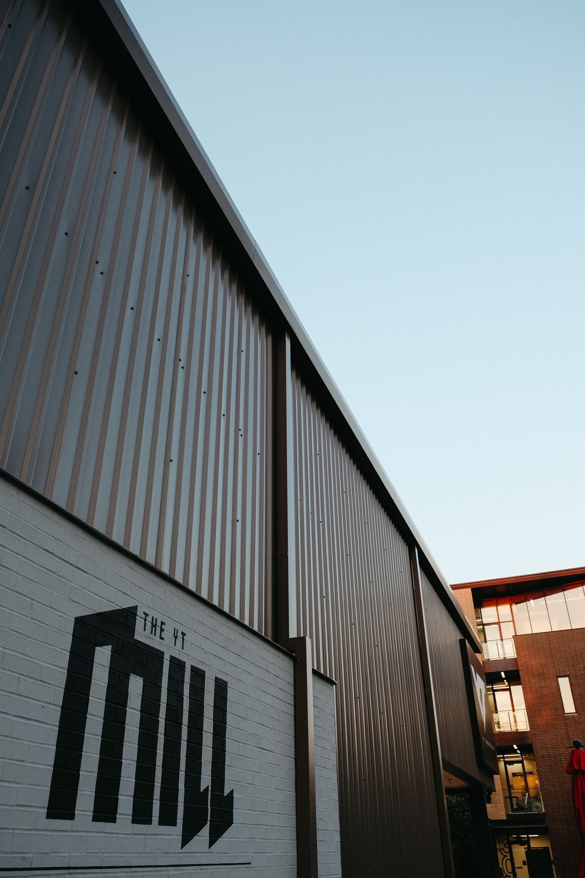 Exterior shot of a modern building with corrugated metal siding and a painted logo on a white wall against a blue sky.