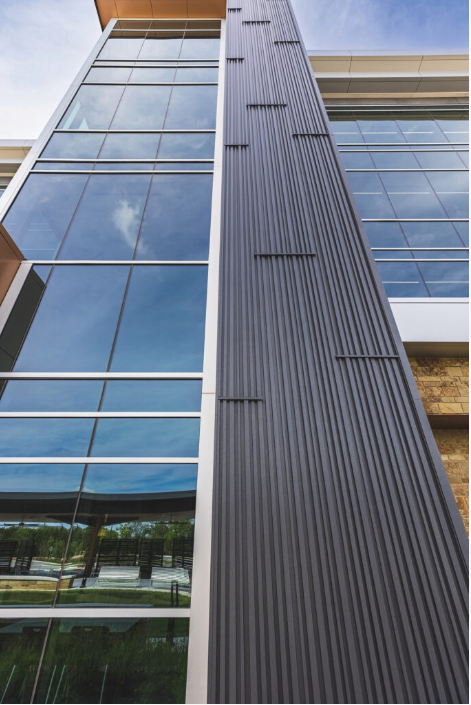 Modern building exterior with dark gray vertical siding, adjacent to glass windows reflecting sky and trees.