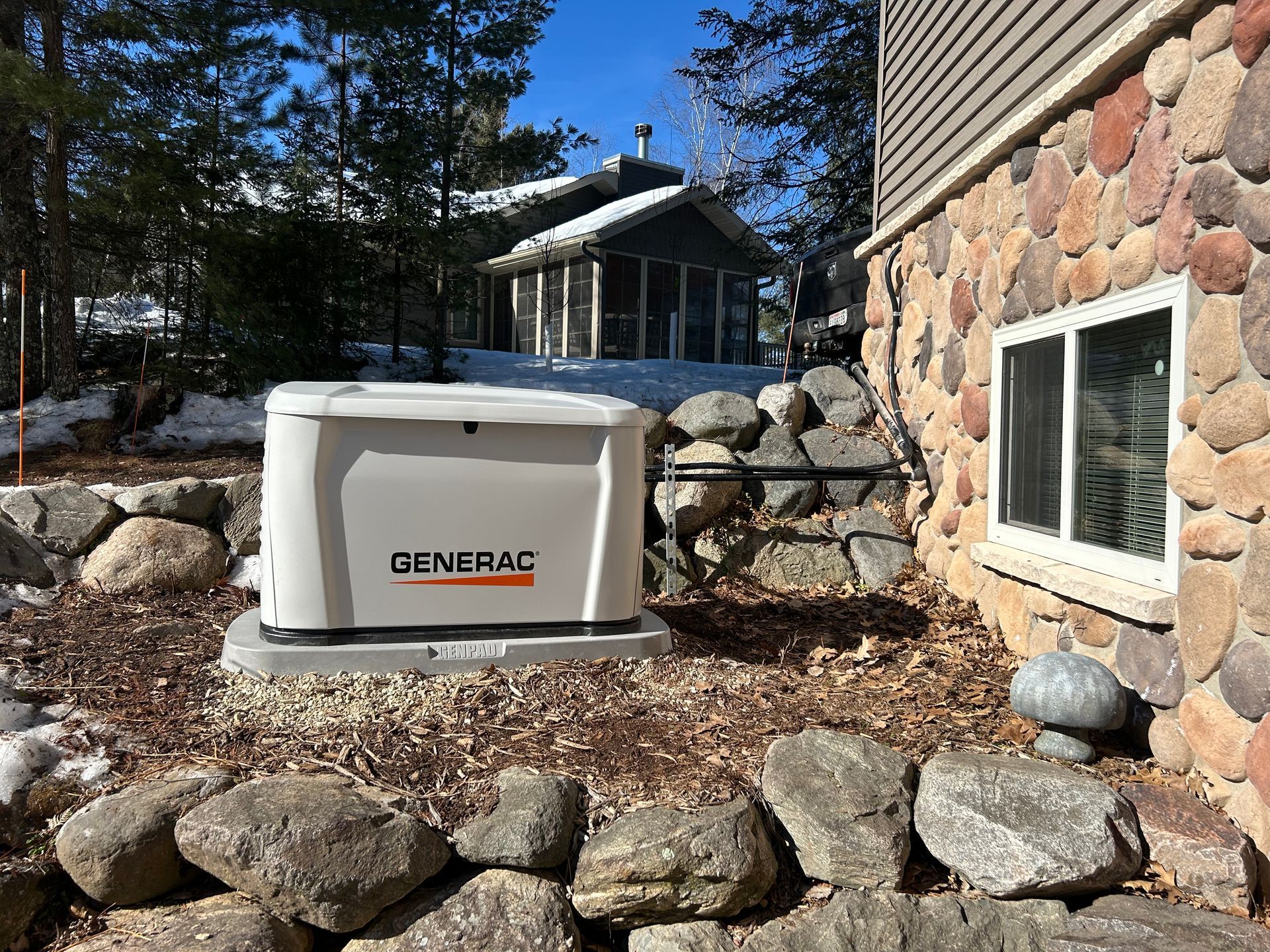 White Generac generator beside a stone house and window, set in a rocky yard with trees and snow.