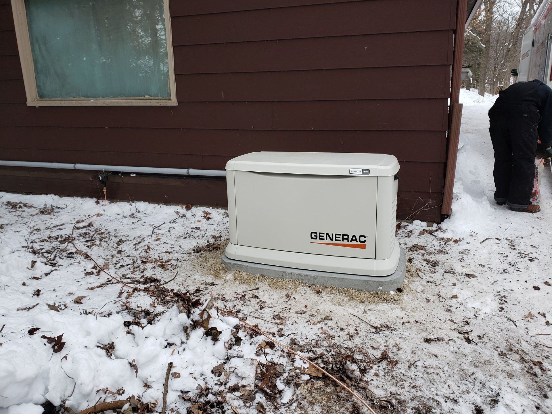 White Generac generator beside a brown house in a snowy yard, with a person clearing snow nearby