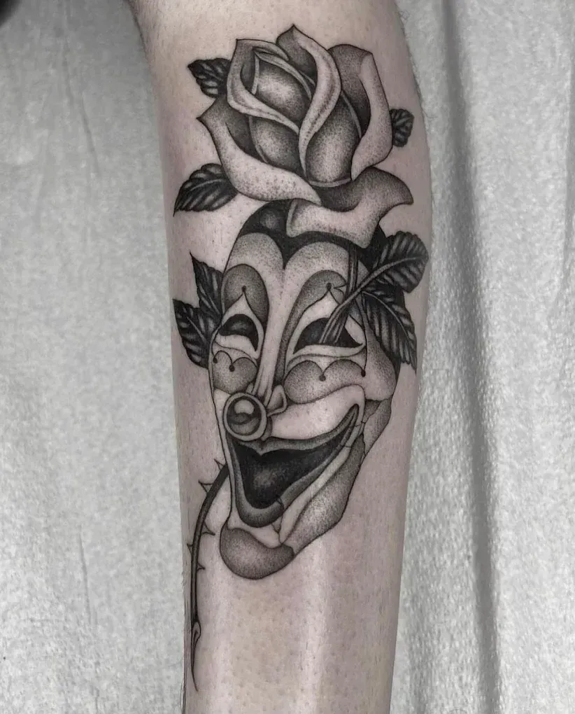 A black and white tattoo of a clown face with a rose.