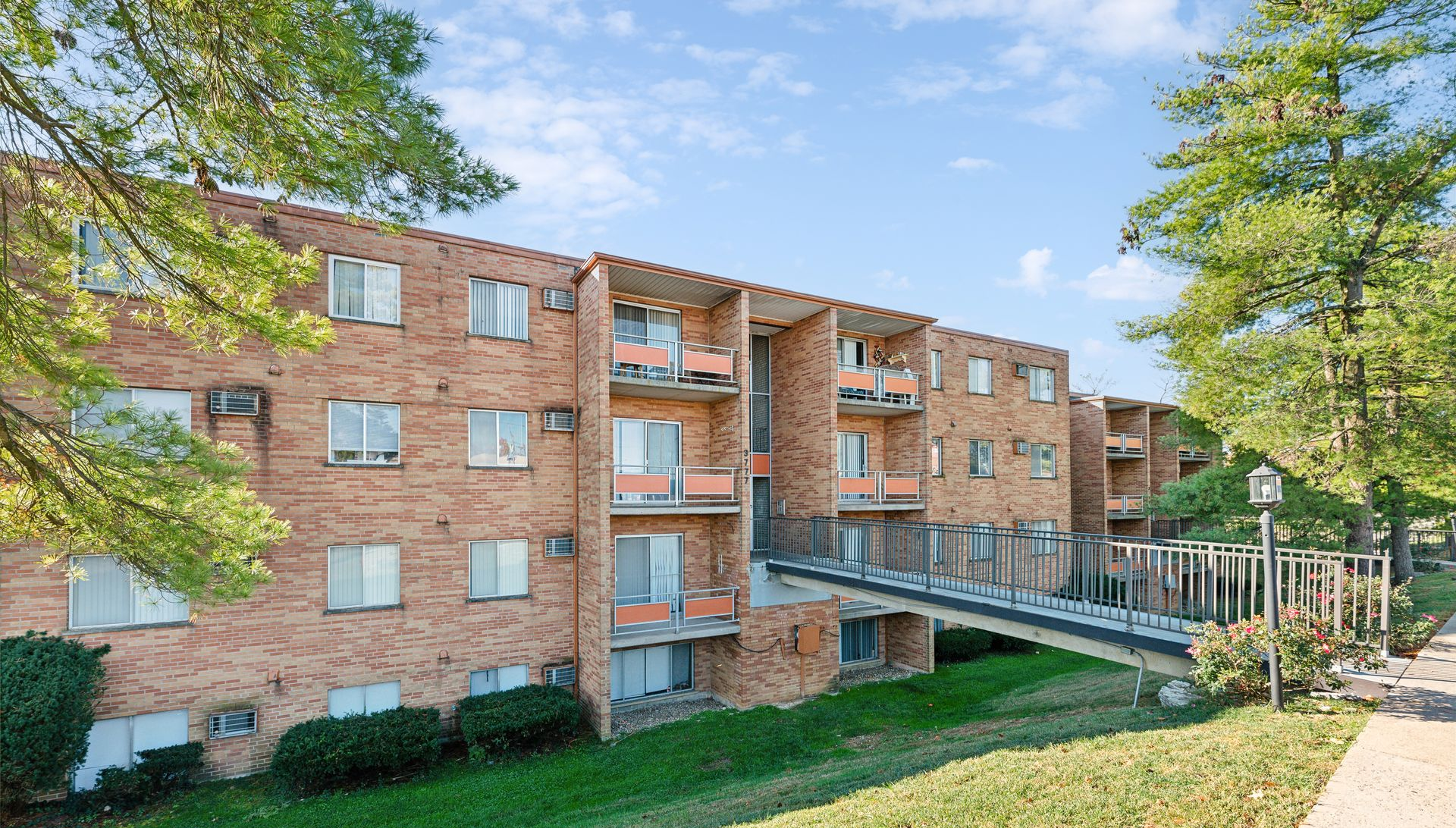Brick apartment building with balconies and a ramp walkway, surrounded by green trees and grass