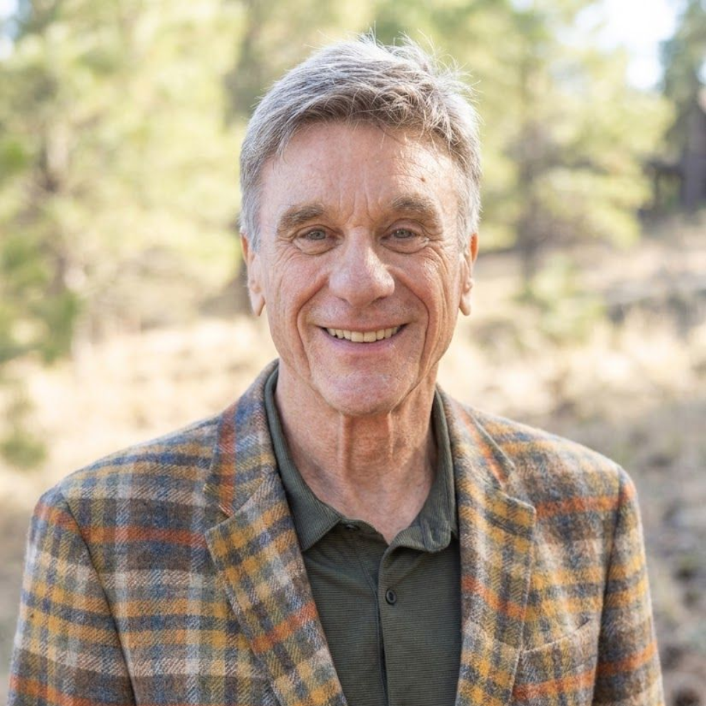 Smiling man in a plaid jacket standing outdoors in a sunlit natural setting