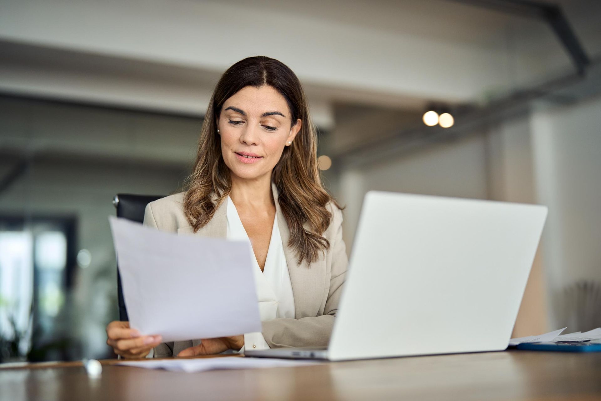 A woman is sitting at a desk with a laptop and holding a piece of paper.