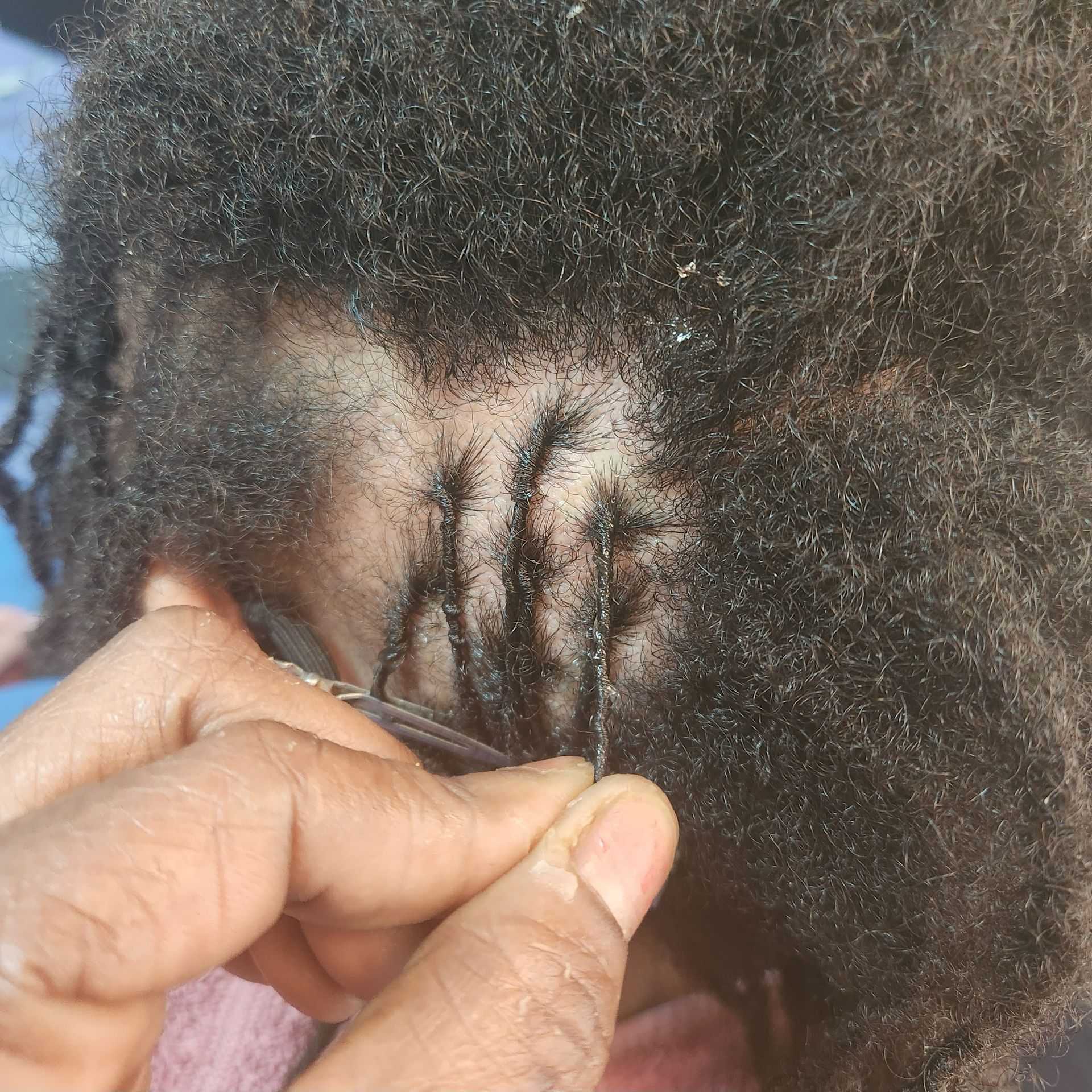 Close-up of dark hair being styled with a tool, likely for loc maintenance. Hands visible.
