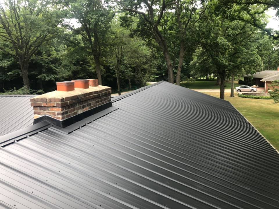 Black metal roof with a brick chimney surrounded by trees and a house in the background. 