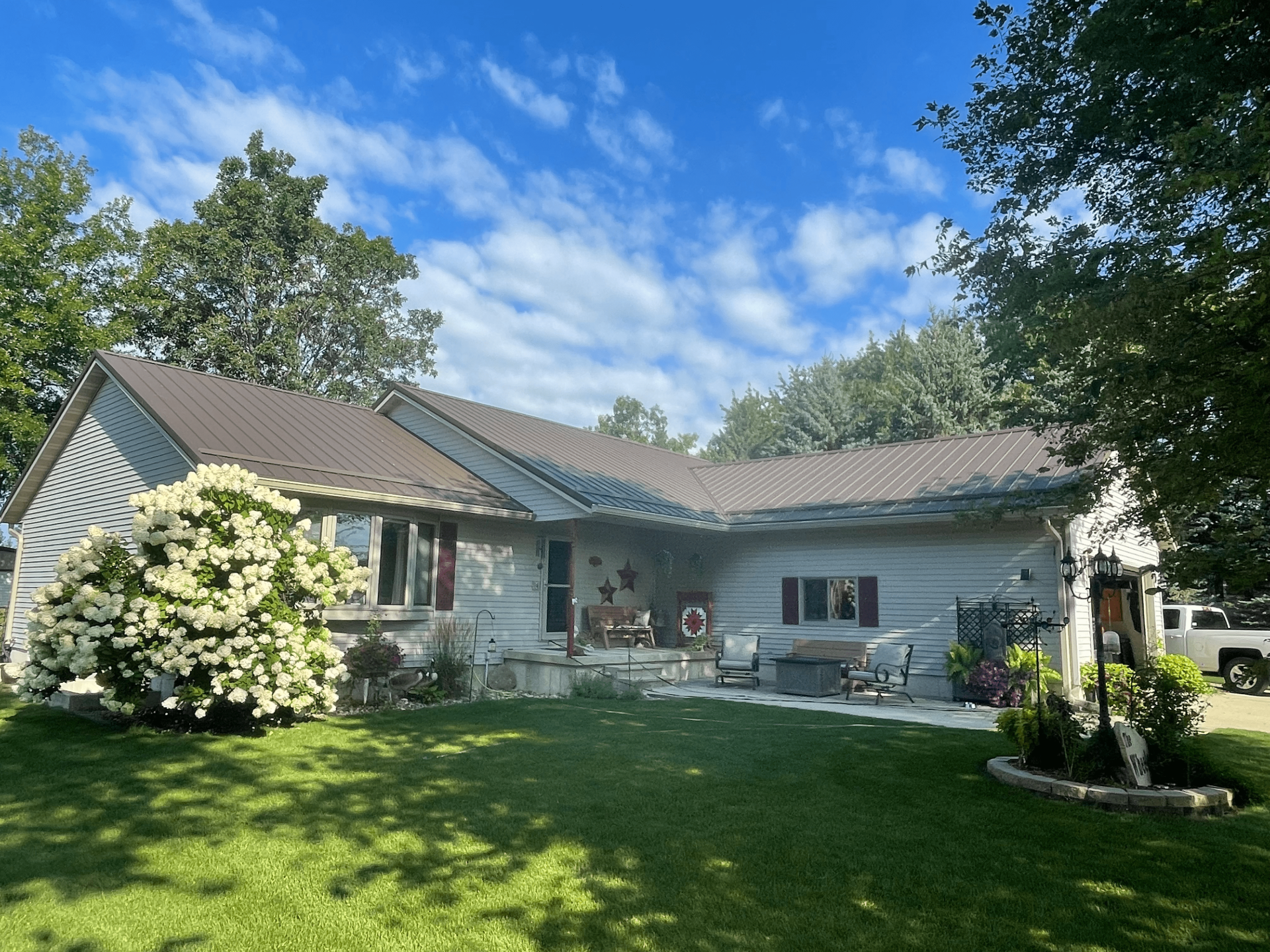 House with brown roof, green lawn, and white and green shrubbery under a blue sky.