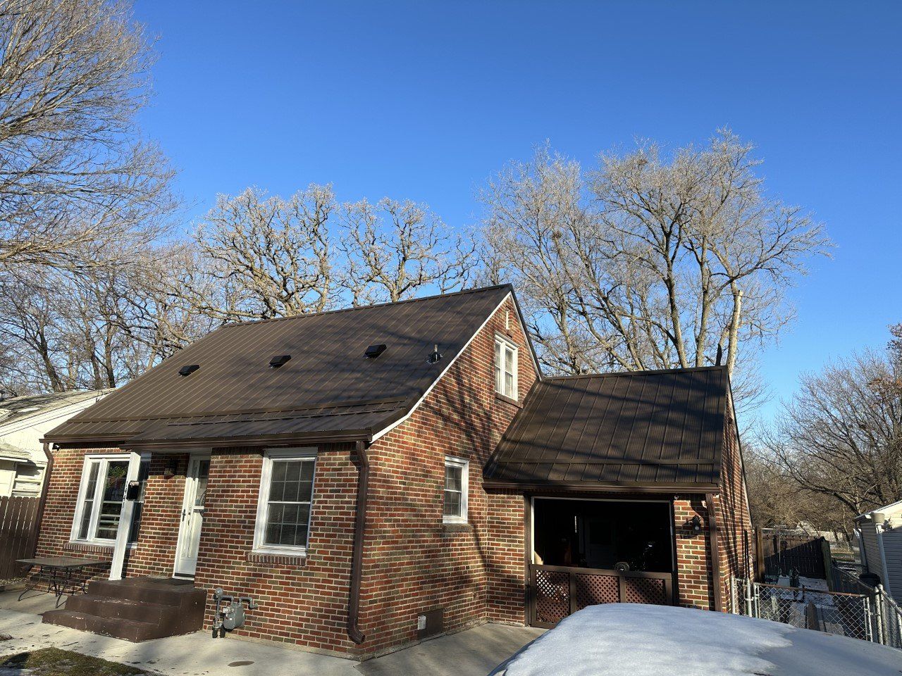 Brick house with brown roof, windows, and snow; trees against blue sky.