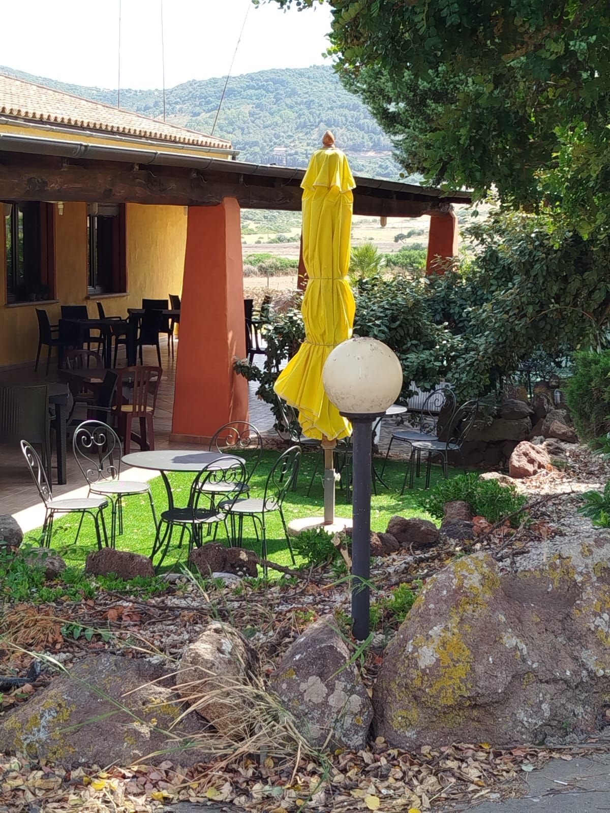 Ombrellone giallo da patio accanto a una serra color pesca, con sedie verdi e aiuole in un giardino soleggiato.