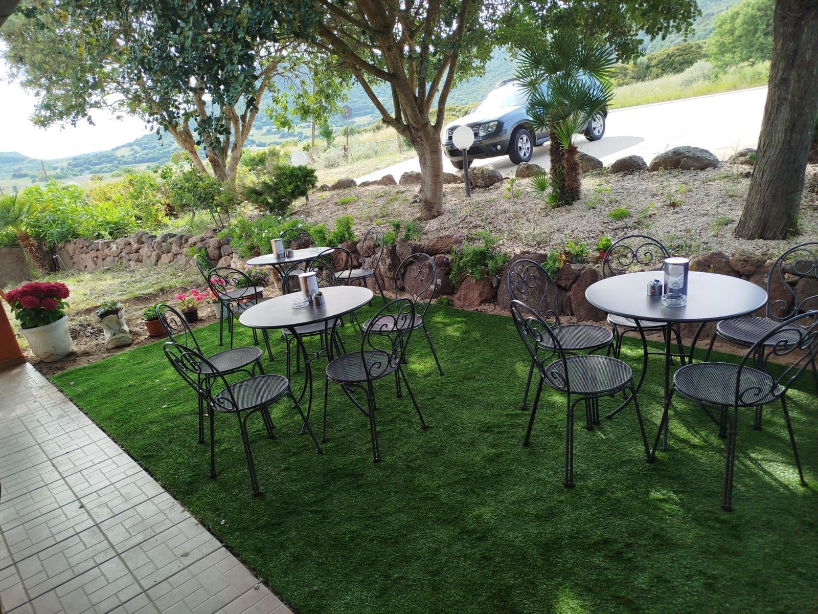 Patio esterno con tavolini rotondi e sedie nere sull'erba, all'ombra degli alberi, con vista su una strada e sulle colline.