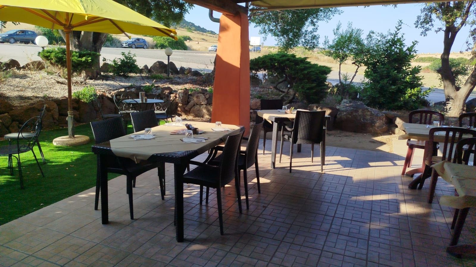Patio esterno con tavoli, sedie e ombrelloni gialli accanto a un giardino in piena giornata