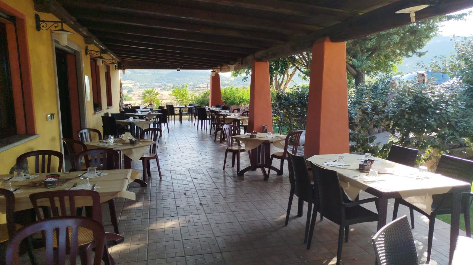 Terrazza esterna coperta del ristorante con tavoli e sedie vuoti, affacciata su un luminoso giardino.