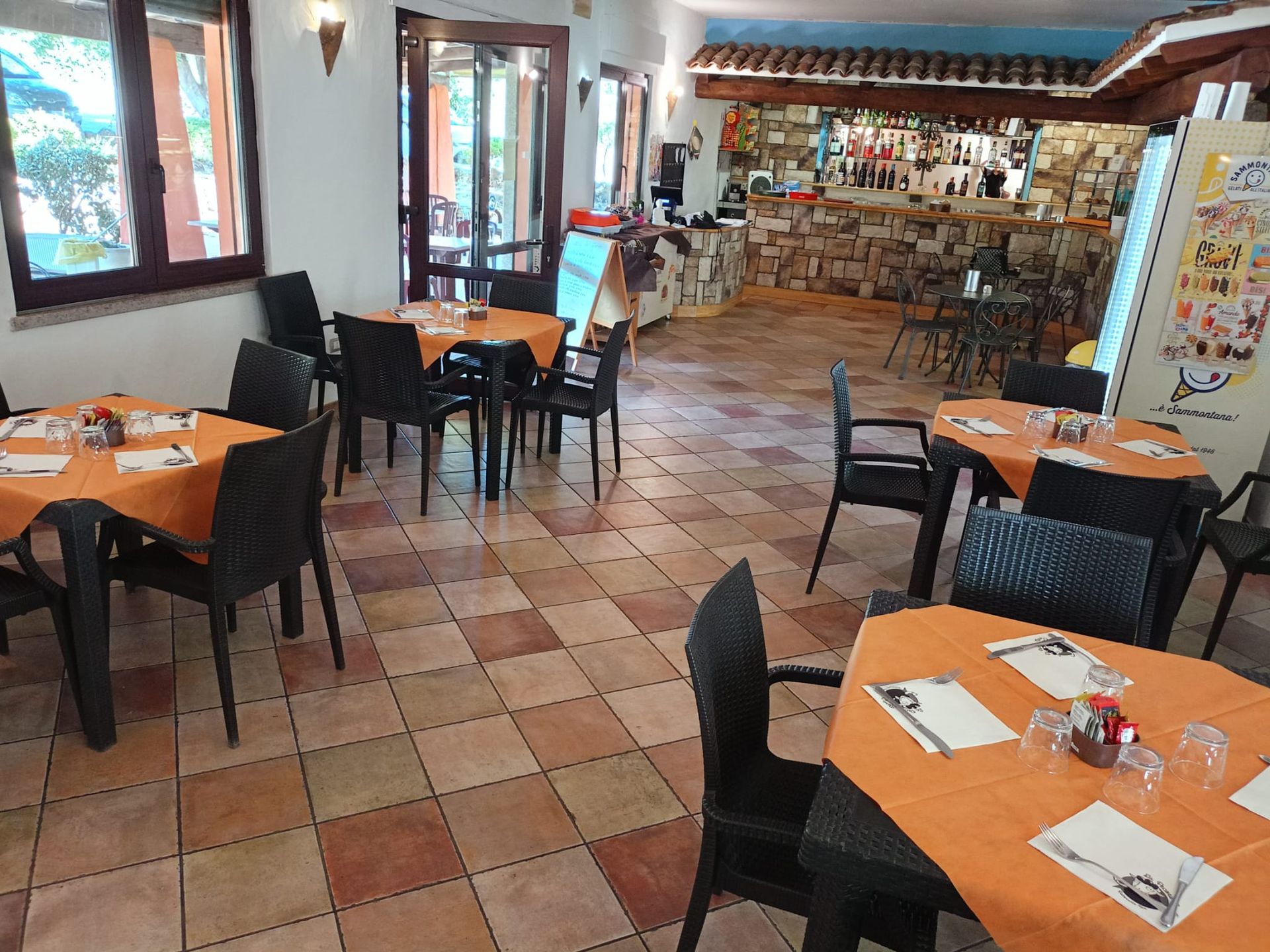 Sala da pranzo vuota di un ristorante con tovaglie arancioni, sedie nere, pavimento piastrellato e un bar sullo sfondo.